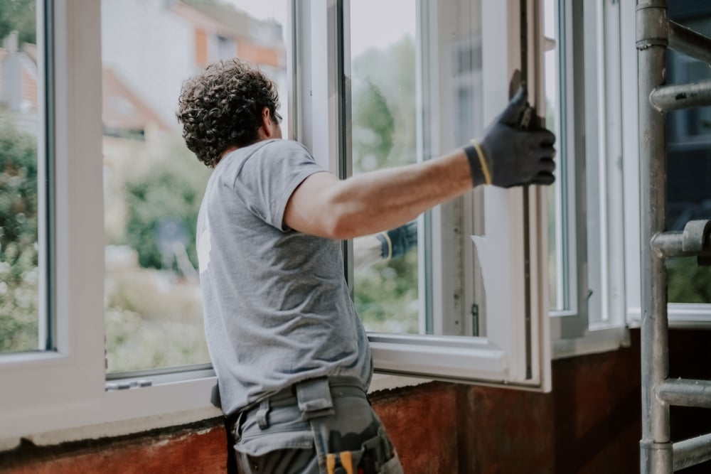 One young handsome Caucasian man in a gray uniform stands from the back and repairs installing a window frame in a house, close-up side view. Concept for home renovation and window installation.