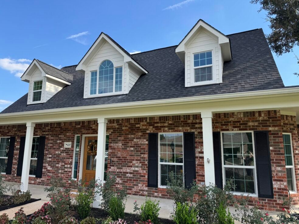 White, brick house facade with dormer windows and a dark shingle roof under a blue sky; front porch and landscaped flower bed in foreground