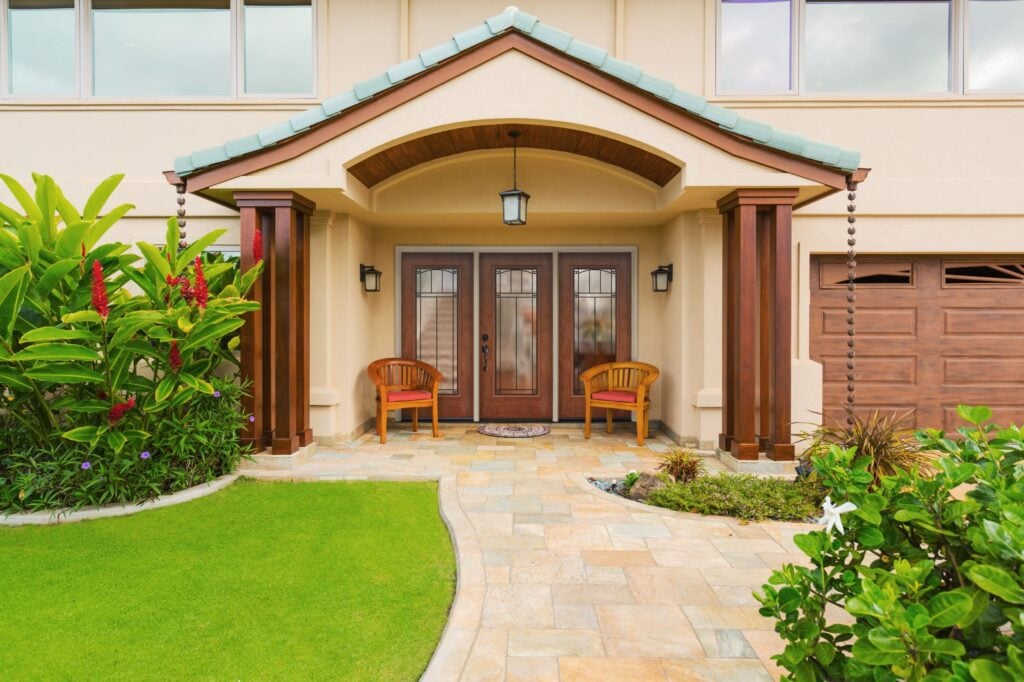 Front entry of a house: double wooden doors with glass, arched porch, two chairs, and a stone path to a green lawn and plants on the side.