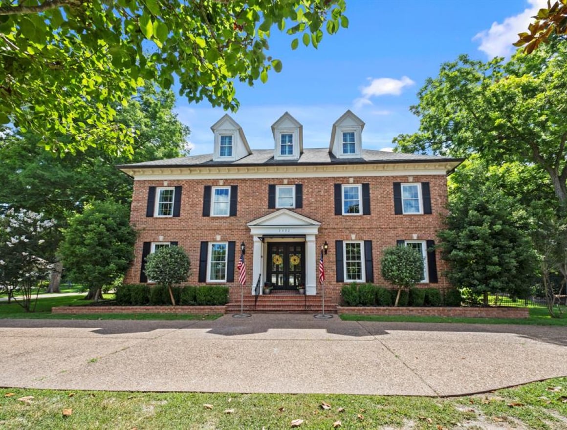 Brick colonial house with dormer windows and a central front door, flanked by two American flags and manicured shrubs on a sunny day