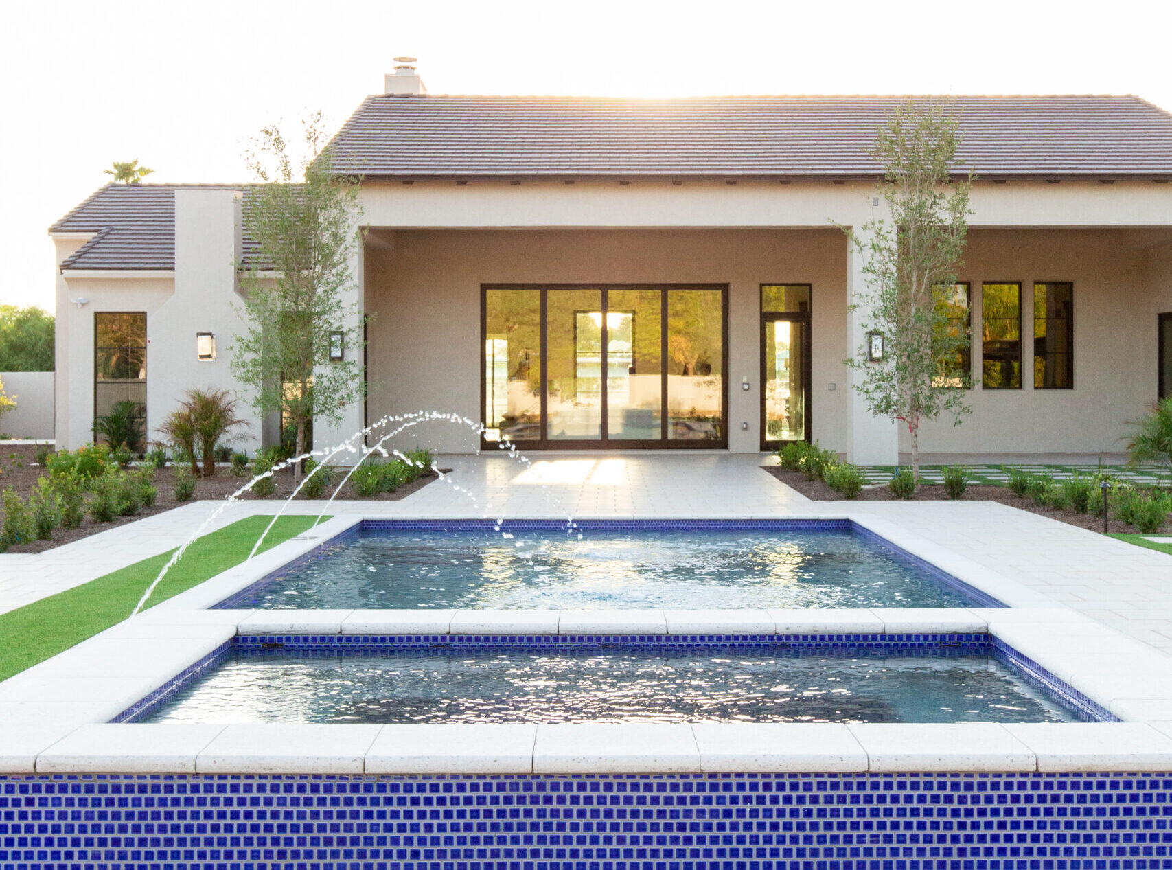 Modern single-story house with large glass doors, front courtyard, and a blue-tiled rectangular pool with fountains in the foreground.