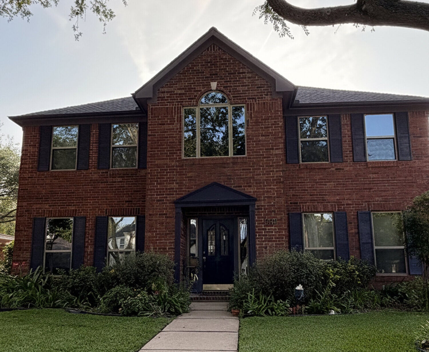 Replacing Vinyl Windows With New Vinyl Windows in Houston Front view of a red brick two-story house with dark shutters, a central arched window above the door, and a concrete walkway leading to the entrance.