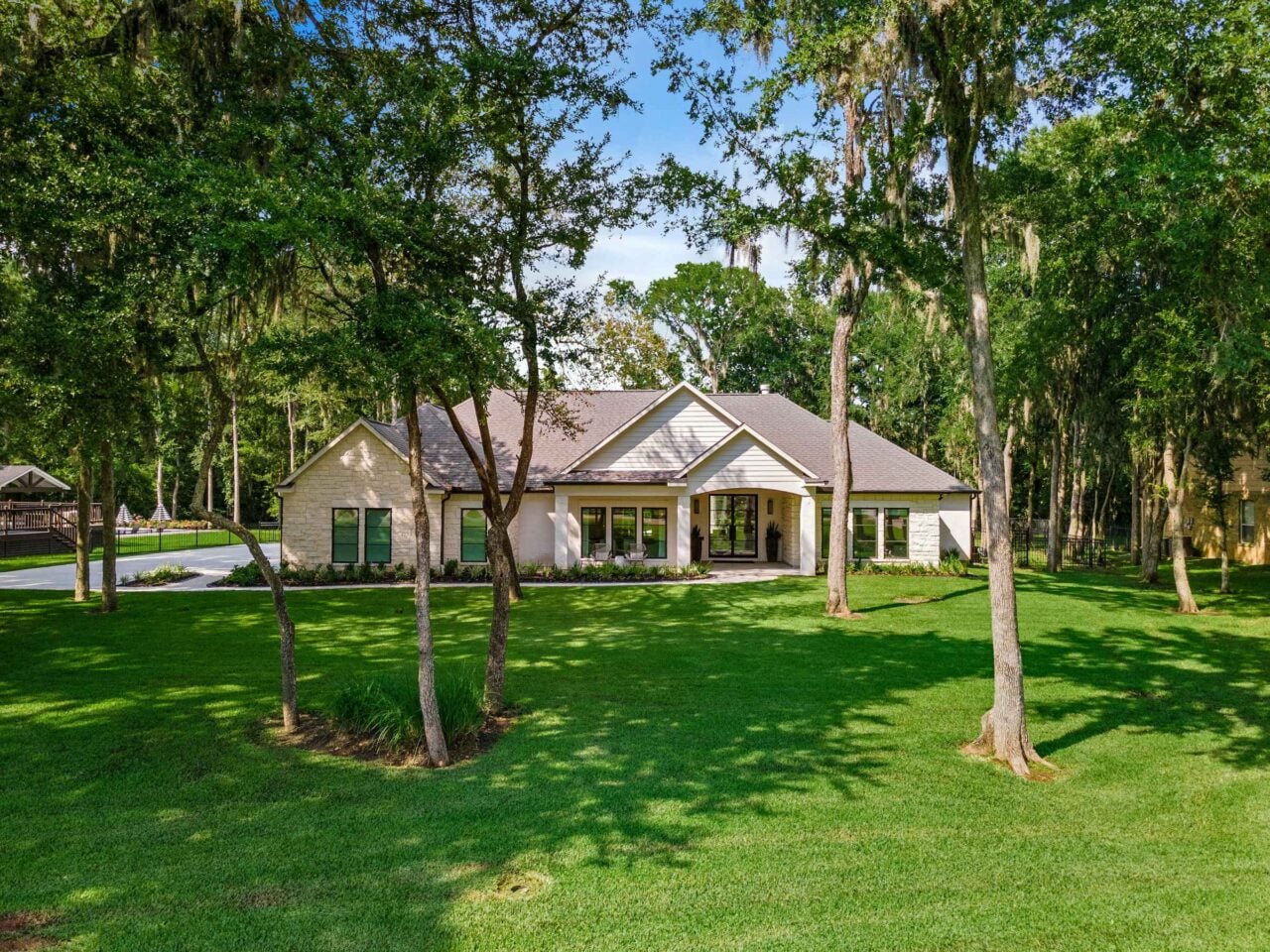 Single-story light-colored house framed by tall trees in a sunny, grassy front yard.
