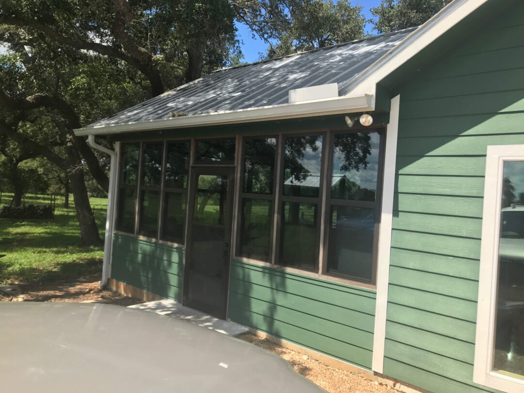Adding More Windows to a House: 5 Key Benefits Green building with large windows, white trim, and a slanted metal roof next to a tree-lined yard, sunny day.