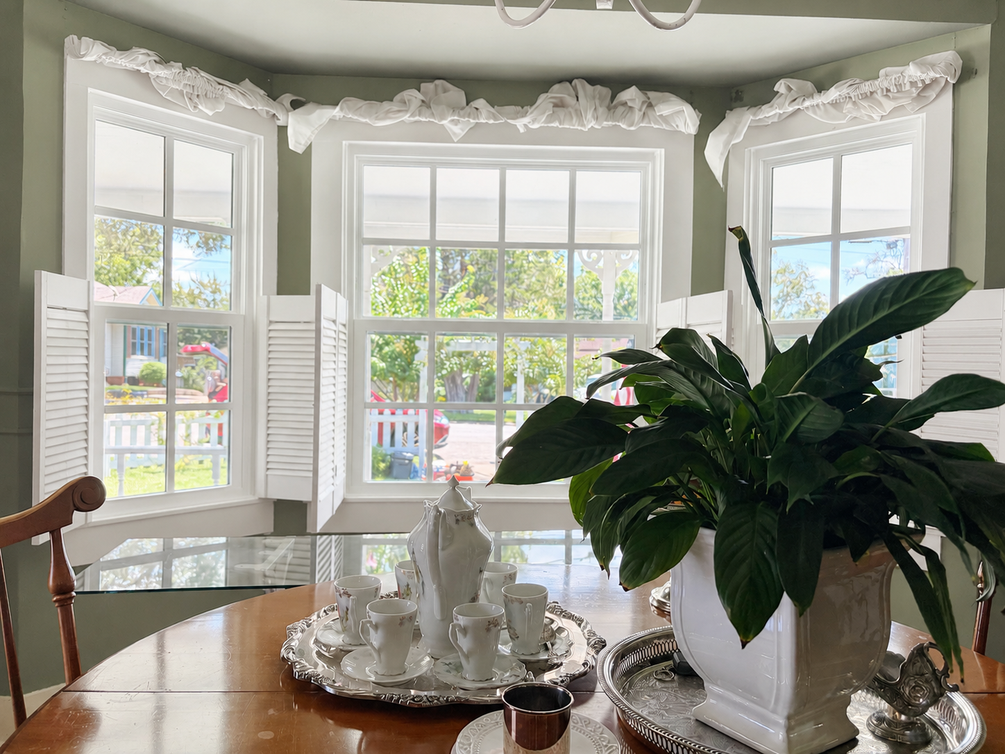 Double Pane Window Glass Replacement: What You Should Know Sunlit dining room with a bay window and white shutters; a large leafy plant on a round wooden table with a tea set on a silver tray nearby.